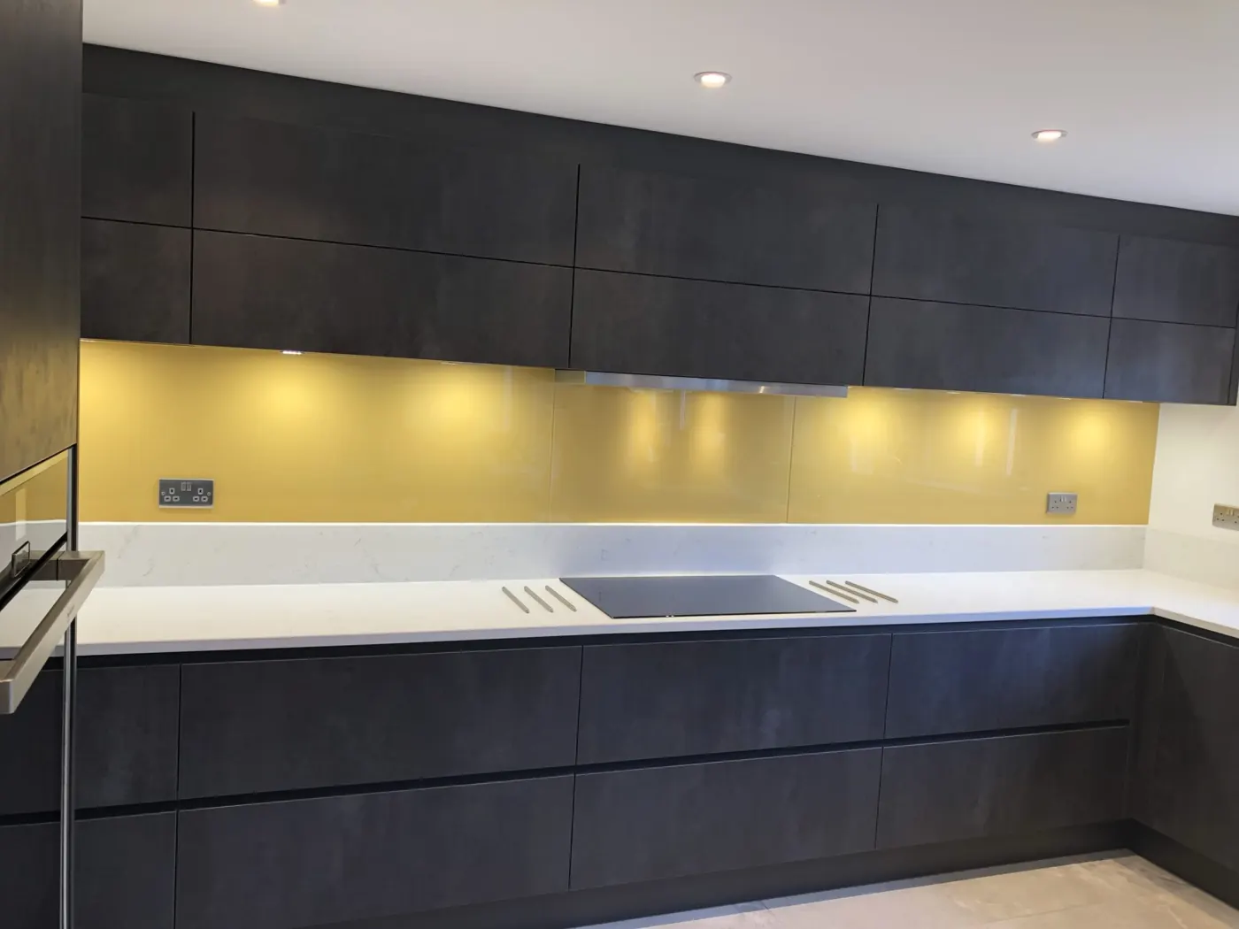 Dark handleless kitchen with a yellow glass splashback and white worktop